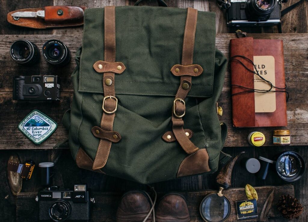 Messer, Kamera, Fotoappart und Film, Rucksack, Schuhe, Lupe, Streichhölzer, Notizbuch, Mückenkreme ausgebreitet auf einem Holztisch
