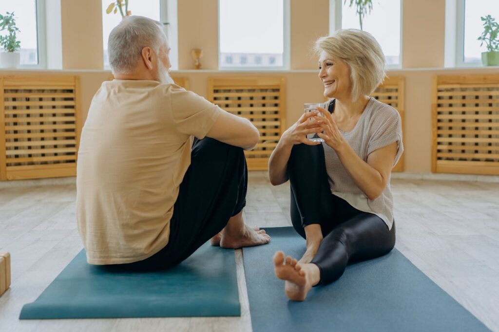 zwei Senioren, Mann und Frau auf Yogamatten sitzend und lächelnd am Unterhalten